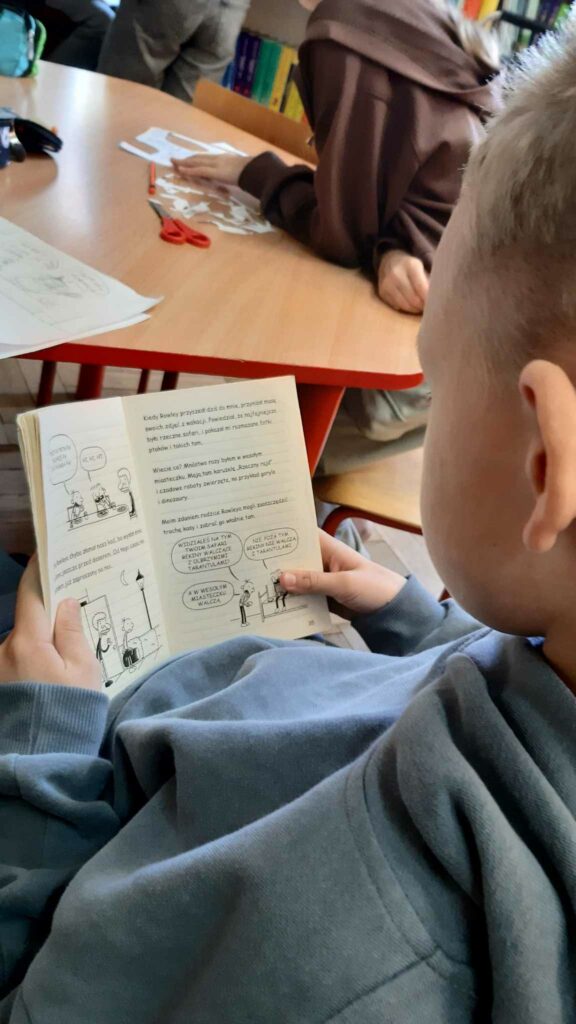 Zdjęcie przedstawia chłopca ustawionego do fotografa tyłem kropka chłopiec trzyma w dłoniach książkę. W tle widać dzieci siedzące przy stole na którym znajduje się pocięta kartka nożyczki ołówek kartki. Chłopiec trzymający książkę ma zdjęcie zrobione w zbliżeniu.