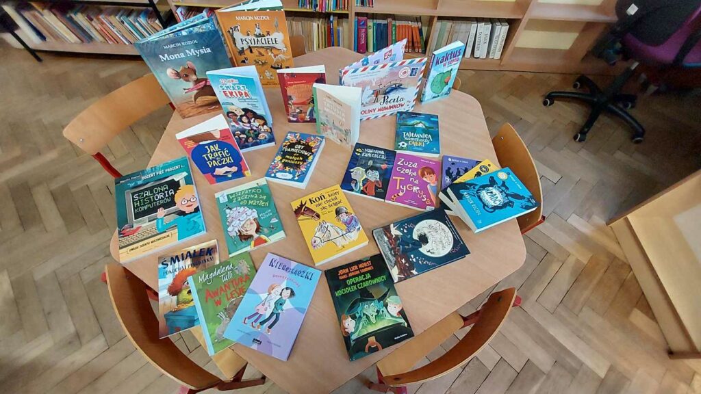 Zdjęcie przedstawia książki ułożone na sześciokątnym stoliku. Wokół stołu stoją krzesełka. Zdjęcie jest zrobione z góry. Książki na stoliku częściowo leżą okładkami z tytułami Górze część książek jest ustawiona tak że widać ich tytuły. W tle widać regały z książkami drewnianą podłogę.