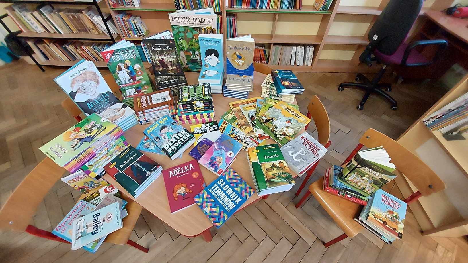 Zdjęcie przedstawia dużą ilość książek ułożoną na sześciokątnym stoliku oraz na krzesełkach obok stolika. Książki są poukładane w stosy lub stoją leżą ułożone w wachlarzu jedna na drugiej. Za książkami widać regały biblioteki.