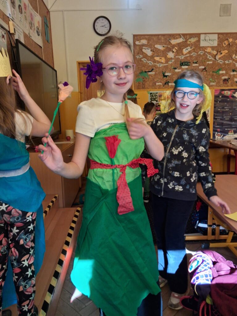 W klasie szkolnej stoją dwie uśmiechnięte dziewczynki. Trzecia stoi odwrócona tyłem do obiektywu. Jedna ma na głowie opaskę z bibuły. Druga ma sukienkę z bibuły, w ręku trzyma kwiat z bibuły. Trzecia dziewczynka ma na głowie koronę z papieru i sukienkę z bibuły.