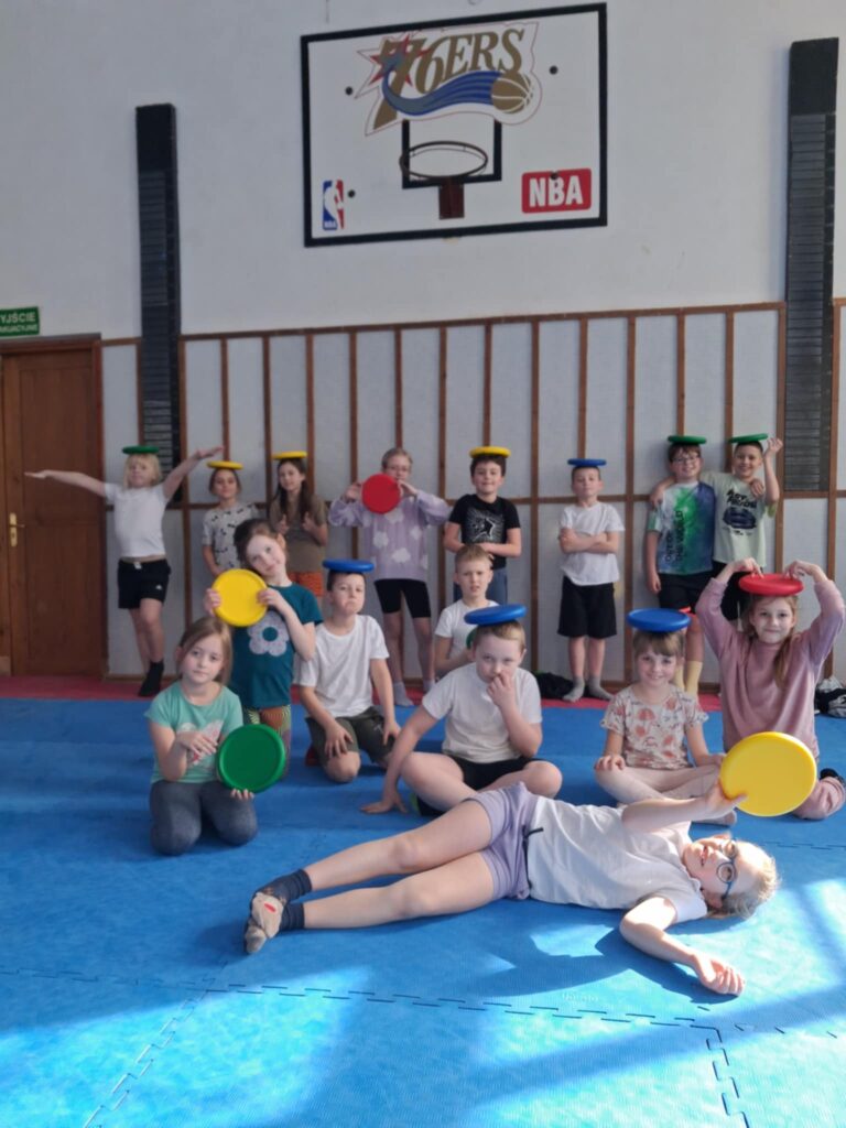 W sali gimnastycznej stoi kilkunastoosobowa grupa dzieci, ubrana w stroje gimnastyczne. Kilkoro dzieci trzyma w rękach kolorowe piankowe freesby. Kilka osób położyło je sobie na głowie. Na pierwszym planie leży dziewczynka, trzyma freesby. Podłoga wyłożona jest niebieską pianką.