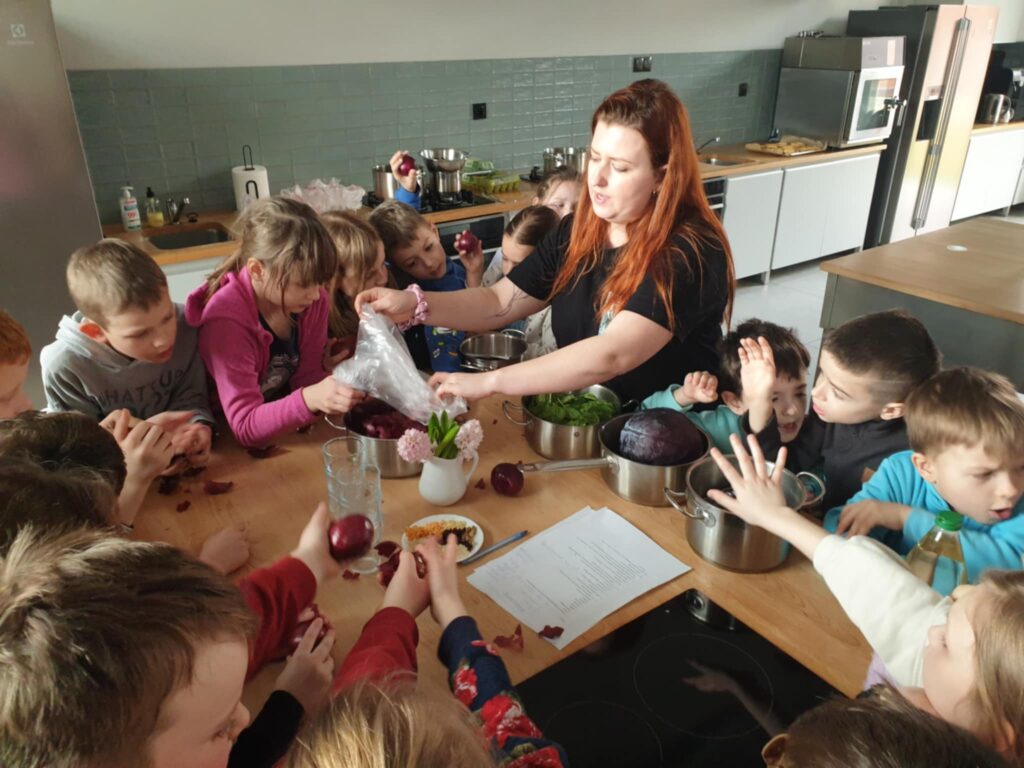 2. Na zdjęciu widać grupę dzieci zgromadzonych wokół stołu kuchennego, gdzie wspólnie z dorosłą kobietą przygotowują naturalne barwniki do barwienia pisanek. Na stole znajdują się garnki z czerwoną kapustą, szpinakiem i cebulą, a dzieci z zaangażowaniem obierają cebulę i sięgają po składniki. Atmosfera jest radosna i pełna entuzjazmu, co wskazuje na twórcze warsztaty wielkanocne.
