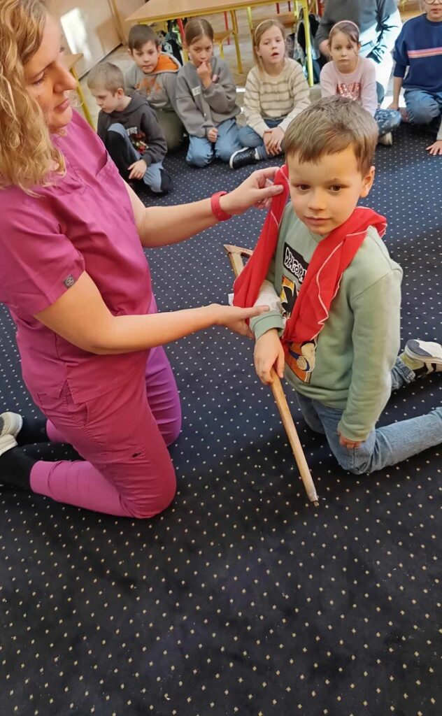 Na zdjęciu widać kobietę w różowym uniformie medycznym klęczącą na dywanie, opatrującą ramię chłopca przy pomocy chusty trójkątnej. Chłopiec ma zieloną bluzę z nadrukiem i trzyma drewniany kij, a jego ręka jest unieruchomiona w temblaku. W tle siedzi grupa dzieci, uważnie obserwujących sytuację, sugerując, że jest to edukacyjna lekcja