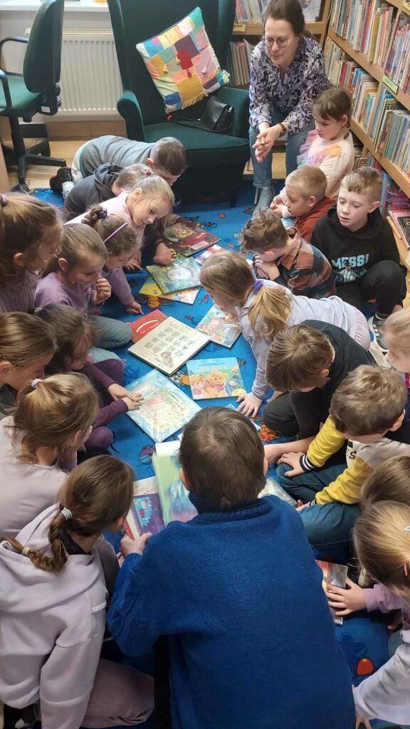 Zdjęcie przedstawia grupę dzieci siedzących na niebieskim dywanie. Dzieci pochylają się nad książkami rozłożonymi pomiędzy nimi. Obok dzieci siedzi kobieta przyglądająca się działaniom dzieci. Obok kobiety stoi duży zielony fotel na którym znajduje się patchworkowa poduszka. W tle widać regały z książkami.