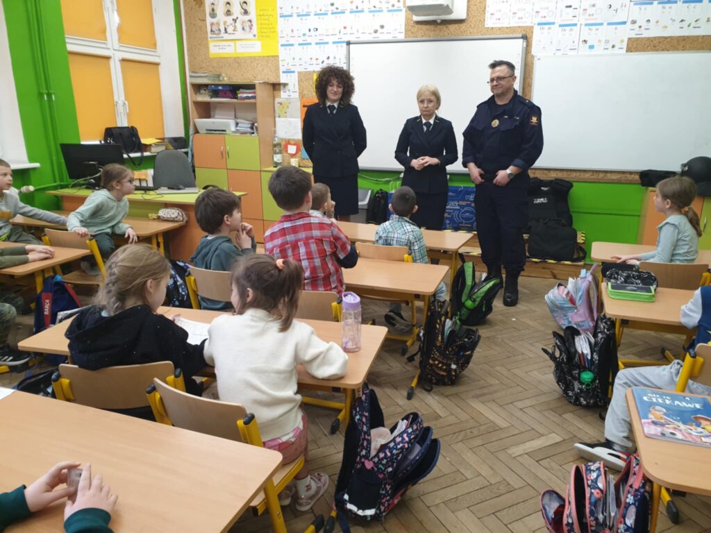 Na zdjęciu widzimy trójkę gości z Kolei Państwowych, którzy odwiedzili klasę szkolną. Ubrani są w służbowe mundury i stoją przed tablicą, rozmawiając z dziećmi siedzącymi w ławkach. Spotkanie ma charakter edukacyjny i dotyczy bezpieczeństwa na kolei.