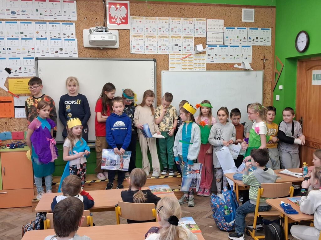 W klasie szkolnej, na tle tablic i plansz z literami, stoi kilkunastoosobowa grupa dzieci. Dzieci są uśmiechnięte. Niektóre z nich mają stroje z bibuły. Dzieci prezentują przedstawienie młodszej klasie.