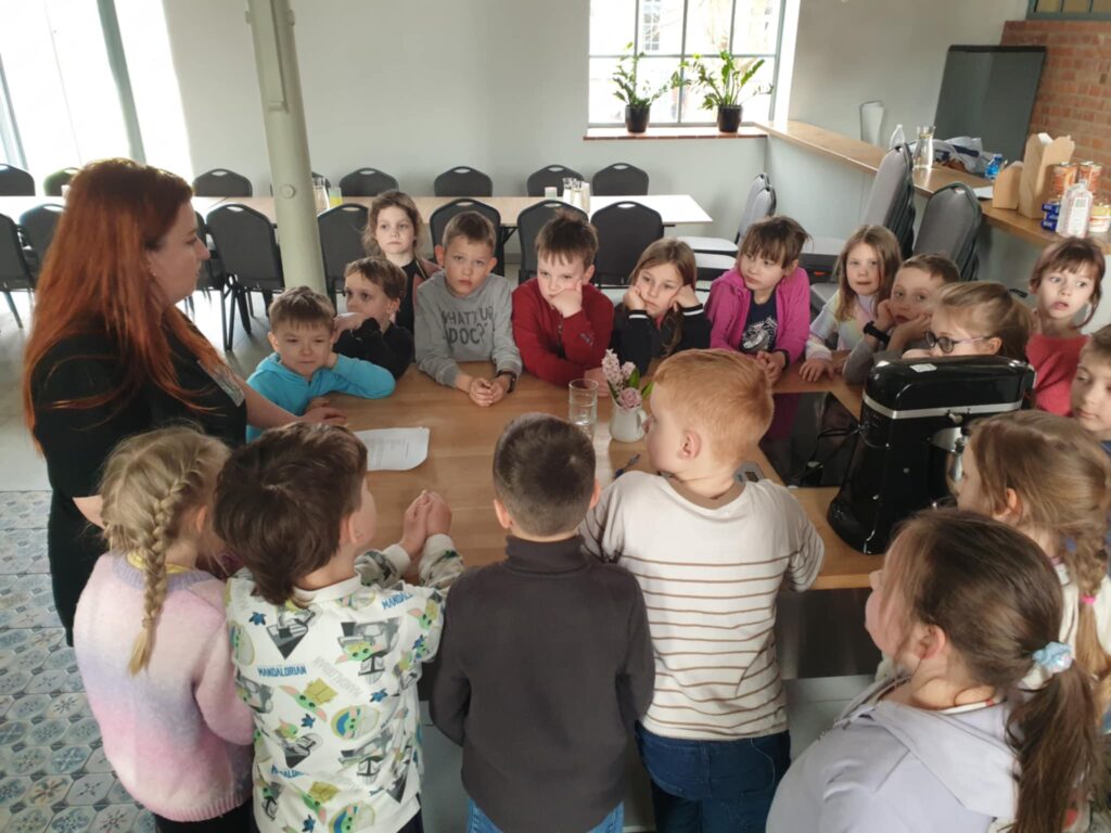 3. Na zdjęciu widzimy grupę dzieci z klasy 1A uczestniczących w warsztatach wielkanocnych. Dzieci zgromadziły się wokół stołu, gdzie prowadząca warsztaty tłumaczy im kolejne kroki zajęć, trzymając kartkę z instrukcjami. W tle widać przytulne wnętrze, a na stole znajdują się różne akcesoria kuchenne, w tym mikser, co sugeruje, że dzieci mogą przygotowywać wielkanocne wypieki.