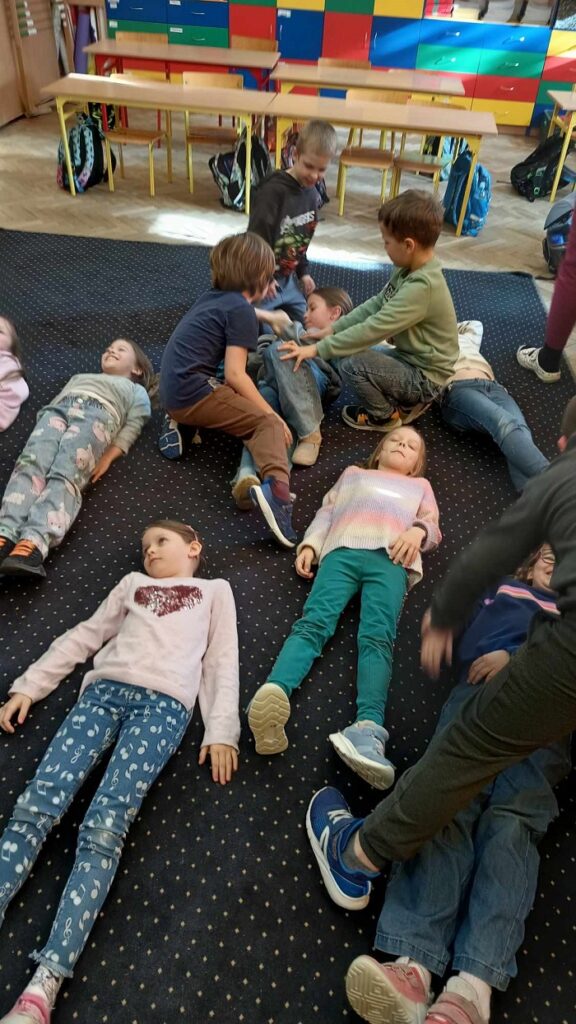 Na zdjęciu znajduje się grupa dzieci bawiących się w sali lekcyjnej, która ma kolorowe szafki i żółte ławki. Niektóre dzieci leżą na ciemnym dywanie, a inne siedzą lub klęczą, jakby uczestniczyły w zabawie lub ćwiczeniu grupowym. W tle widać plecaki zawieszone na krzesłach oraz jasną podłogę z parkietem.