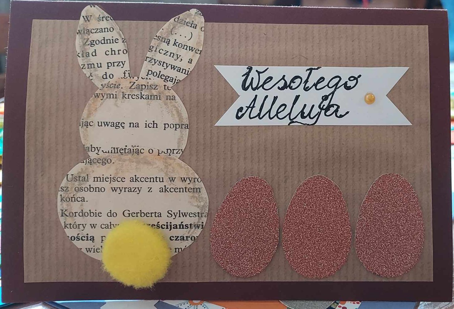 Zdjęcie przedstawia kartke świąteczną z napisem Wesołego Alleluja zająca ustawionego tyłem z widocznym żółtym ogonkiem oraz trzy jajka papierowe