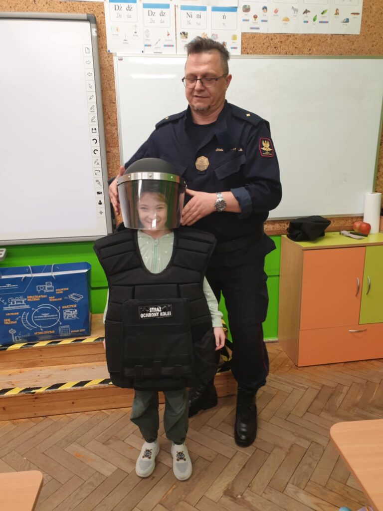 Na zdjęciu widzimy funkcjonariusza Straży Ochrony Kolei w ciemnym mundurze, który pomaga dziecku założyć hełm ochronny oraz kamizelkę kuloodporną. Dziewczynka o imieniu Kaja, uśmiecha się, przymierzając sprzęt ochronny, który jest na nią zdecydowanie za duży. Zdjęcie zostało wykonane w klasie szkolnej, bo była to edukacyjna wizyta funkcjonariuszy SOK w szkole.