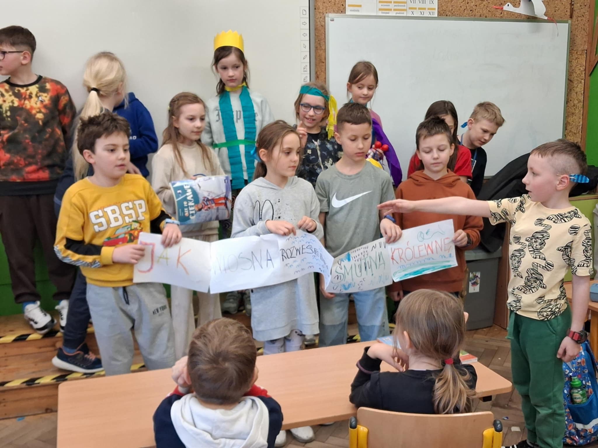 Cały kadr zajmuje grupa dzieci. Na pierwzsym planie stoją 4 osoby i trzymają listwę z napisem. Po prawej stronie stoi chłopiec, który wskazuje ręka napis. Za nimi widać dzieci, część z nich ma elementy stroju z papieru i bibuły.