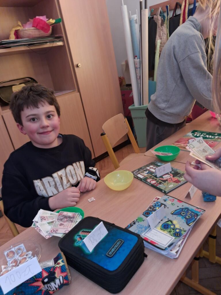 Na zdjęciu widać fragment ławki szkolnej. po lewj stronie siedzi za nią uśmiechnięty chłopiec. po parwej stronie stoi za nia chłopiec, który układa przedmioty na ławce. Chłopcy przygotowali sklepik klasowy. Na ławce widać różne przedmioty, kartki z cenami, papierowe pieniądze. W tle widać regał z książkami.