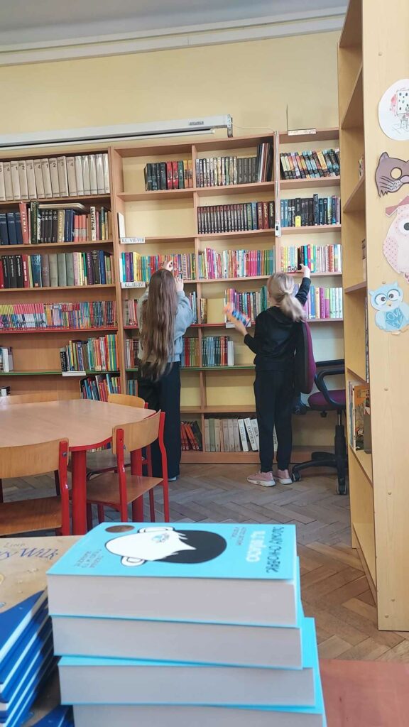 Zdjęcie przedstawia dwie dziewczynki stojące przodem do regałów z książkami. Dziewczynki przeglądają znajdujące się na regałach książki. Na pierwszym planie widać książki poukładane w stos kropka Na zdjęciu widać stolik z kilkoma krzesełkami wokół.