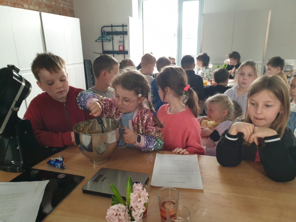 4. Na zdjęciu widać grupę dzieci w kuchni, które biorą udział w warsztatach kulinarnych. Na pierwszym planie dziewczynka w błyszczącej kurtce miesza ciasto w metalowej misce, a inne dzieci uważnie się temu przyglądają. warsztaty