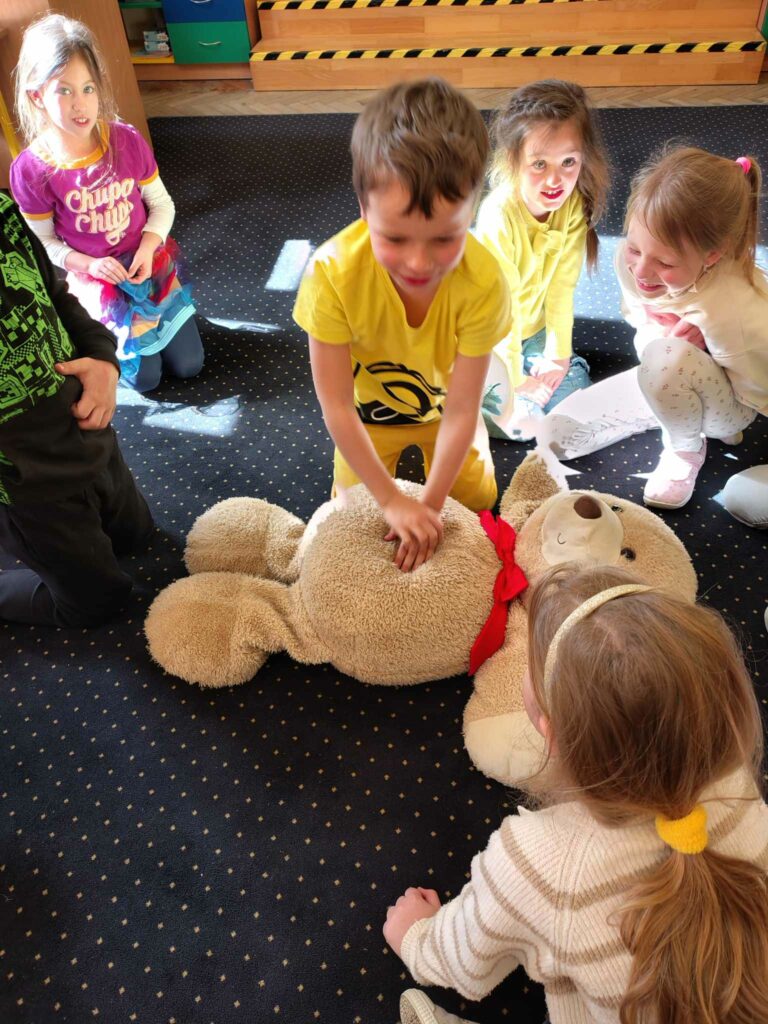 Na zdjęciu widać grupę dzieci bawiących się w symulację udzielania pierwszej pomocy dużemu pluszowemu misiowi. Chłopiec w żółtej koszulce wykonuje uciski na klatce piersiowej misia, podczas gdy reszta dzieci z zainteresowaniem obserwuje. Dzieci są uśmiechnięte i zaangażowane, co sugeruje, że zajęcia mają charakter edukacyjny i zabawowy jednocześnie.