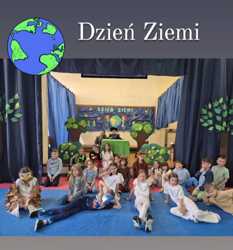 Na zdjęciu znajduje się duża grupa dzieci przebranych na zwierzęta leśne oraz ubranych na biało i niebiesko. Dzieci siedzą na podłodze na sali gimnastycznej. W tle widać dekoracje: papierowe drzewa oraz tablicę z napisem "dzień ziemi".