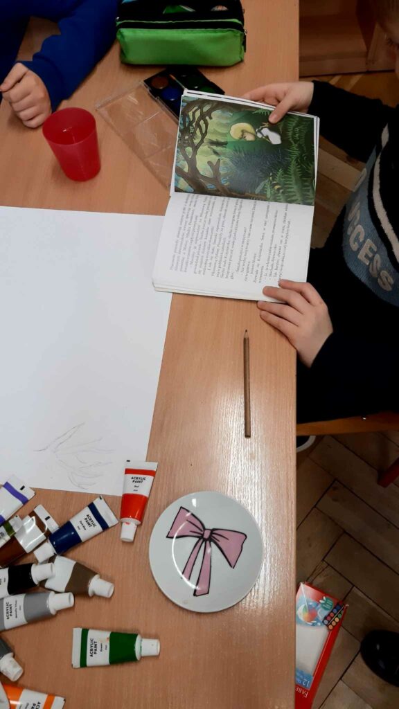 Zdjęcie przedstawia dzieci siedzące przy ławce. Na ławce znajdują się farby kartka papieru talerzyk z grafiką kokardki kredka kubek z wodą kropka Dzieci mają oparte ręce o ławkę jedno dziecko trzyma książkę otwartą.