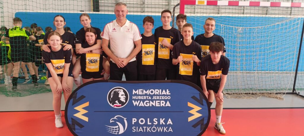 Zdjęcie przedstawia drużynę siatkarską, prawdopodobnie młodzieżową, pozostającą w zwartym gronie z trenerem. Zawodnicy i trener trzymają w rękach żółte transparenty z napisem "MEMORIAL 2024 WAGNER". Stoją przed banerem reklamowym z logo "MEMORIAL HUBERTA JERZEGO WAGNERA" oraz logo Polskiej Siatkówki. Uczestnicy ubrani są w czarne koszulki z tym samym napisem, a trener w białą bluzę. Tło stanowi hala sportowa z widoczną siatką do siatkówki. Atmosfera jest oficjalna i radosna, sugerująca udział w turnieju lub ważnym wydarzeniu siatkarskim. Zdjęcie jest ostre i dobrze naświetlone, co pozwala na wyraźne rozpoznanie szczegółów.