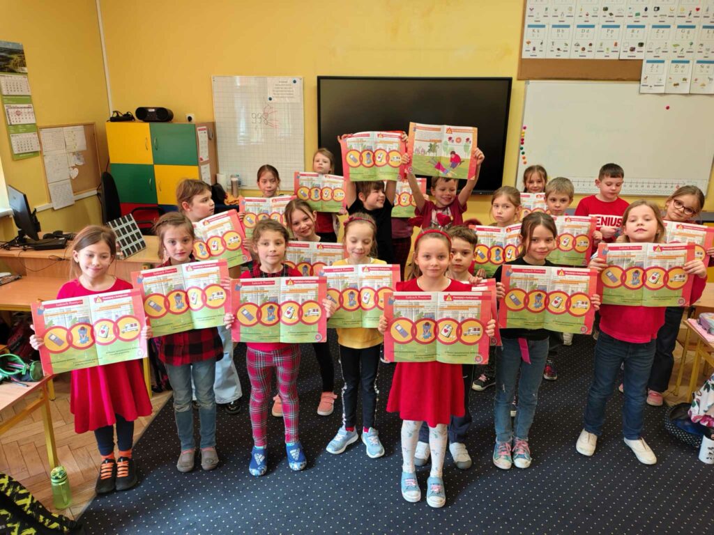 Na zdjęciu znajduje się grupa dzieci stojących w klasie szkolnej i trzymających w rękach kolorowe broszury edukacyjne. Dzieci są uśmiechnięte i ubrane w czerwone lub czerwono-czarne ubrania. W tle widać wyposażenie klasy, takie jak tablica, regały, komputery oraz materiały dydaktyczne.