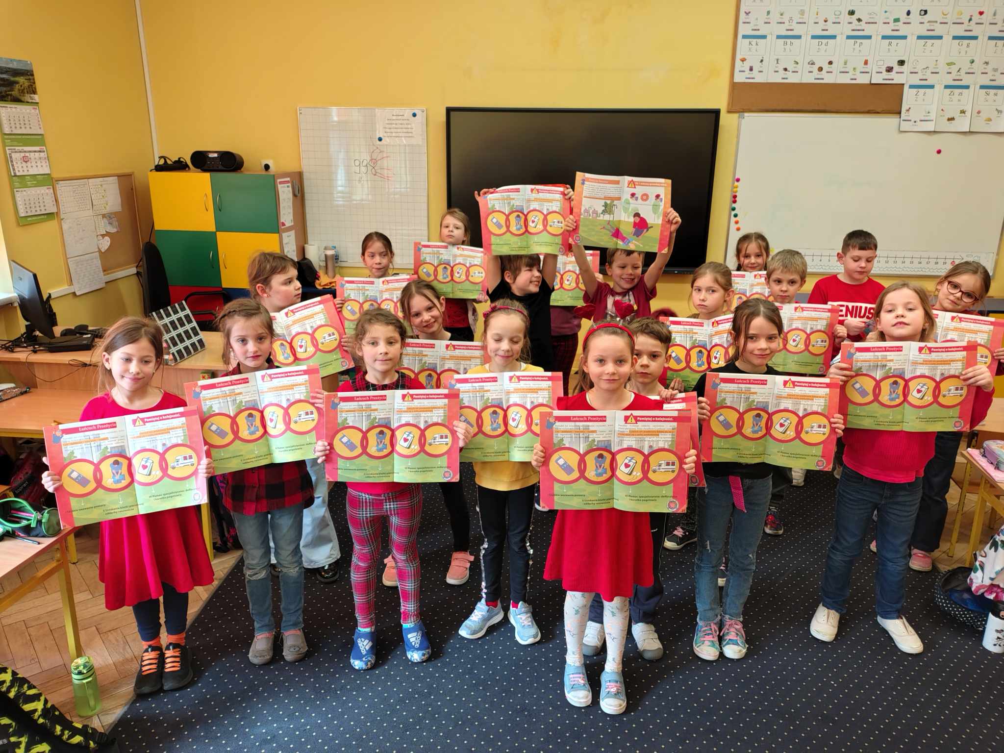 Na zdjęciu znajduje się grupa dzieci stojących w klasie szkolnej i trzymających w rękach kolorowe broszury edukacyjne. Dzieci są uśmiechnięte i ubrane w czerwone lub czerwono-czarne ubrania. W tle widać wyposażenie klasy, takie jak tablica, regały, komputery oraz materiały dydaktyczne.