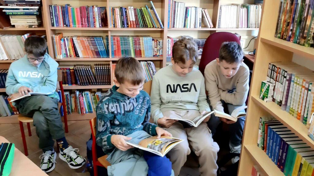 Zdjęcie przedstawia czterech chłopców siedzących na na krzesełkach. Chłopcy trzymają w rękach książki. Czytają. W tle widać regały z książkami.