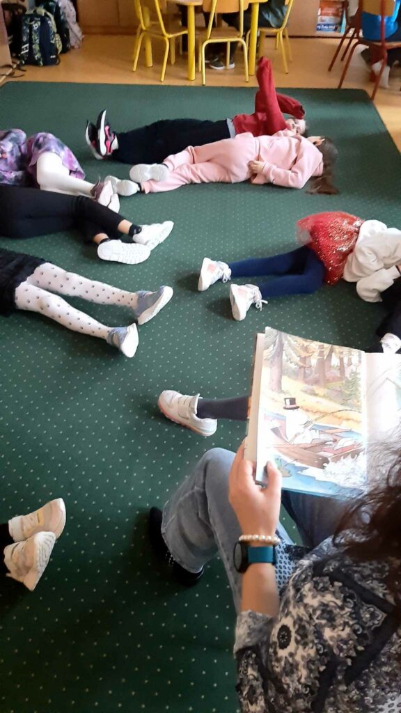 Zdjęcie przedstawia dzieci leżące na zielonym dywanie. Niektóre dzieci mają podniesione ręce do góry u innych widać tylko nogi z prawej strony fotografii widać częściowo kobietę trzymającą w dłoni książka.