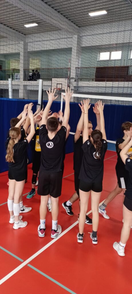 Zdjęcie przedstawia grupę młodych ludzi, prawdopodobnie uczniów, którzy radośnie świętują sukces na boisku do siatkówki. Ubrani są w jednakowe czarne koszulki i krótkie spodenki, a ich białe skarpetki i buty sportowe podkreślają sportowy charakter chwili. Wszyscy podnoszą ręce w geście triumfu, tworząc zwarty krąg na tle siatki do siatkówki i części trybun. Atmosfera jest pełna energii i pozytywnych emocji, sugerując wspólny wysiłek i osiągnięcie wspólnego celu. Jasne, ostre zdjęcie idealnie ukazuje radość i entuzjazm młodych sportowców. Scena jest dynamiczna i pełna życia, a uśmiechy na twarzach (choć niewidoczne, można je wywnioskować z gestu) sugerują niezapomniane przeżycie.