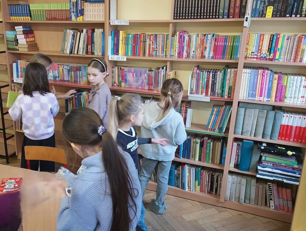 Zdjęcie przedstawia sześć dziewczynek które stoją przy regałach z książkami. Dziewczynki wybierają książki. Część z nich jest ustawiona do fotografa tyłem część bokiem.