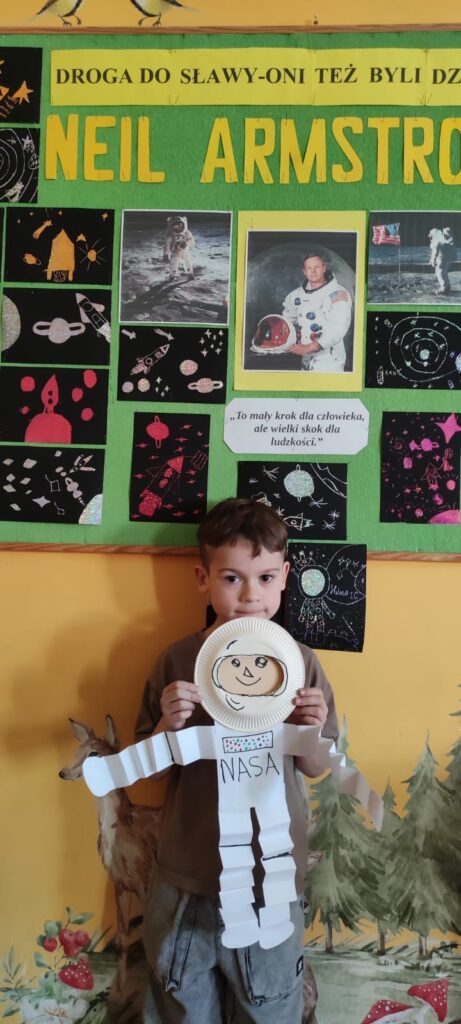 Na zdjęciu widzimy chłopca stojącego na tle kolorowej gazetki szkolnej poświęconej Neilowi Armstrongowi – pierwszemu człowiekowi, który stanął na Księżycu. Gazetka zawiera jego zdjęcia, prace plastyczne dzieci przedstawiające rakiety i kosmos oraz cytat: „To mały krok dla człowieka, ale wielki skok dla ludzkości.” Chłopiec trzyma w rękach własnoręcznie wykonaną postać astronauty z papierowego talerzyka i białych pasków papieru, podpisaną „NASA”. Astronauta ma narysowaną twarz, hełm oraz detale kombinezonu. Całość wygląda na projekt edukacyjny poświęcony eksploracji kosmosu i postaci Neila Armstronga. Tło ściany to przyjazny mural z motywami leśnymi – widoczne są drzewa, lis oraz grzyby. Atmosfera zdjęcia jest radosna i edukacyjna.