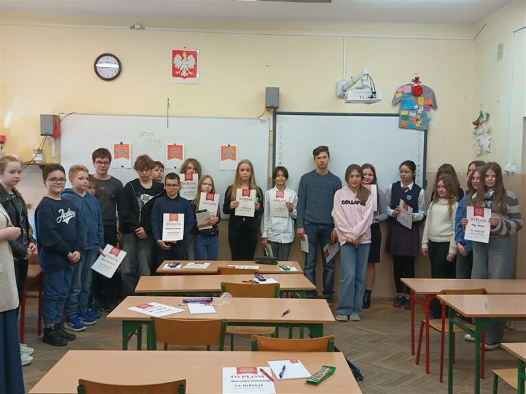 na zdjęciu widać większą grupę uczniów, którzy również stoją w klasie. W tle widoczna jest tablica, zegar oraz godło Polski. Uczniowie prezentują swoje dyplomy, a na stolikach widać dodatkowe nagrody. Atmosfera jest uroczysta,