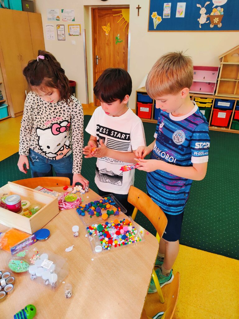 Na zdjęciu troje dzieci stoi przy stole i wspólnie tworzy prace plastyczne. Na stole leżą różnorodne materiały kreatywne, takie jak kolorowe pompony, naklejki, farby i oczka. Dzieci są skupione na swojej pracy i wydają się zaangażowane w twórczą zabawę. W tle widać salę przedszkolną z kolorowymi szafkami i dekoracjami świątecznymi na tablicy.