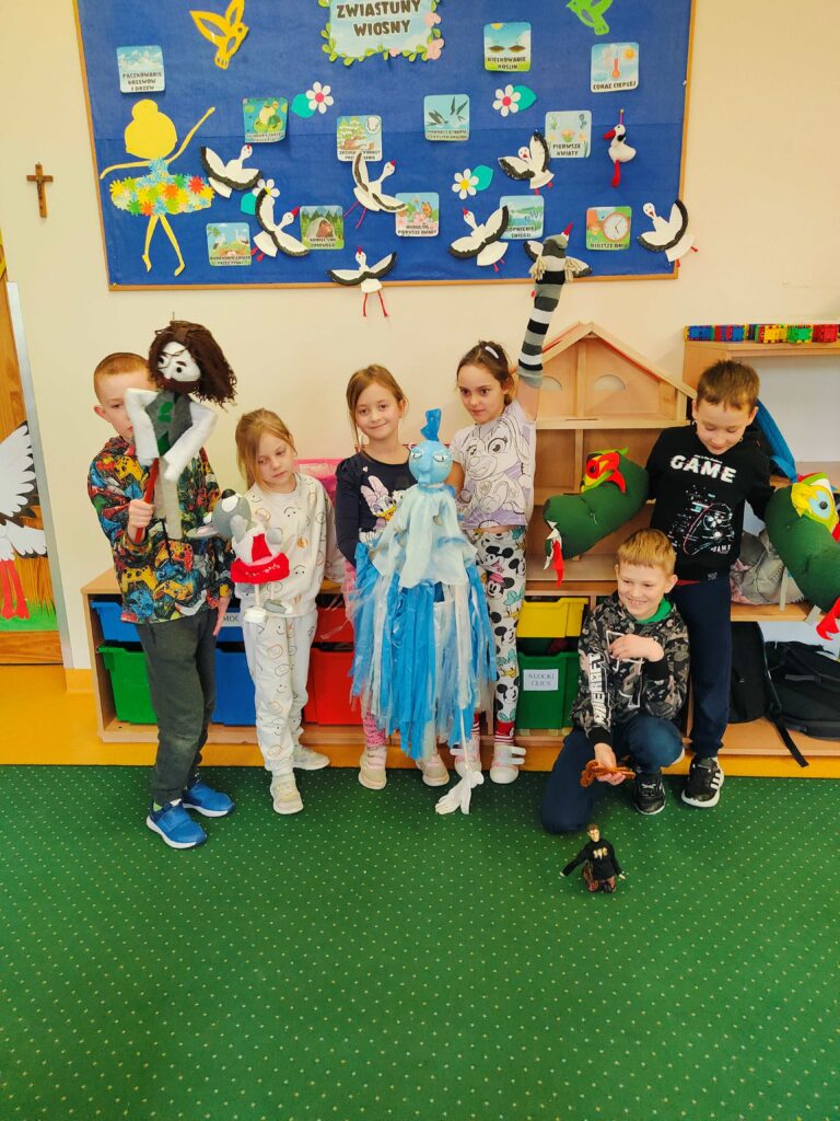 Na zdjęciu znajduje się grupa sześciu dzieci stojących w klasie, trzymających kukiełki i lalki teatralne. W tle widać tablicę z napisem „Zwiastuny wiosny”, udekorowaną rysunkami i papierowymi bocianami. Dzieci mają na sobie kolorowe ubrania i wyglądają na zadowolone, prezentując swoje prace. Klasa jest przytulnie urządzona, z półkami pełnymi kolorowych pudełek.