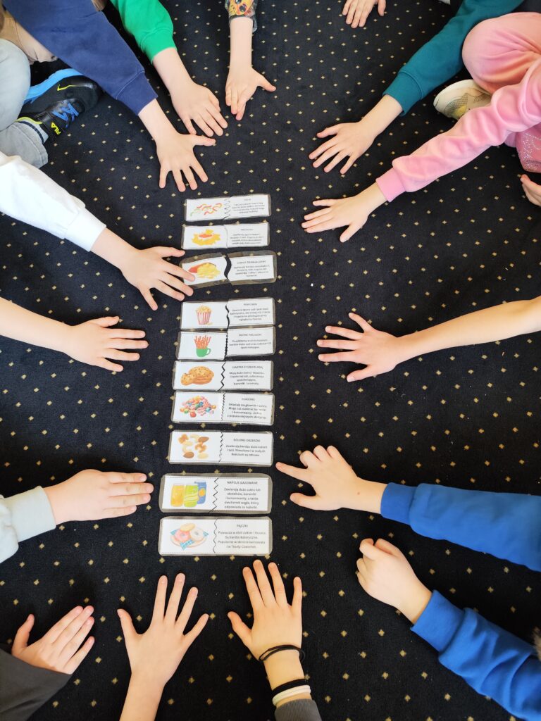Na zdjęciu widać dzieci siedzące na podłodze w kręgu, wyciągające ręce w stronę kart z informacjami o różnych produktach spożywczych. Karty przedstawiają ilustracje i opisy żywności, takiej jak frytki, chipsy, słodkie napoje czy ciastka. Podłoga jest pokryta ciemnym dywanem w jasne kropki. Dzieci są zaangażowane w edukacyjną aktywność, dotyczącą zdrowego odżywiania.