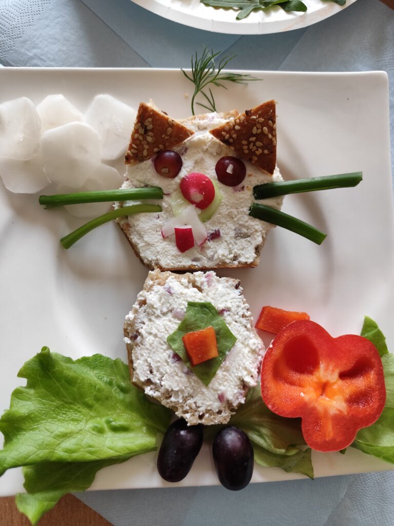 Na zdjęciu widzimy kreatywną kanapkę w kształcie kota wykonaną z pieczywa, twarożku i warzyw. Głowa zwierzaka to kromka chleba posmarowana twarogiem, a uszy zostały zrobione z trójkątnych kawałków chleba z ziarnami. Oczy są wykonane z winogron, nos z rzodkiewki, a wąsy ze szczypiorku. Buzię zdobią drobne kawałki rzodkiewki i ogórka. Całość została ułożona na białym talerzu, a obok znajdują się dodatkowe elementy dekoracyjne, takie jak plasterki białej rzodkwi oraz gałązka koperku.