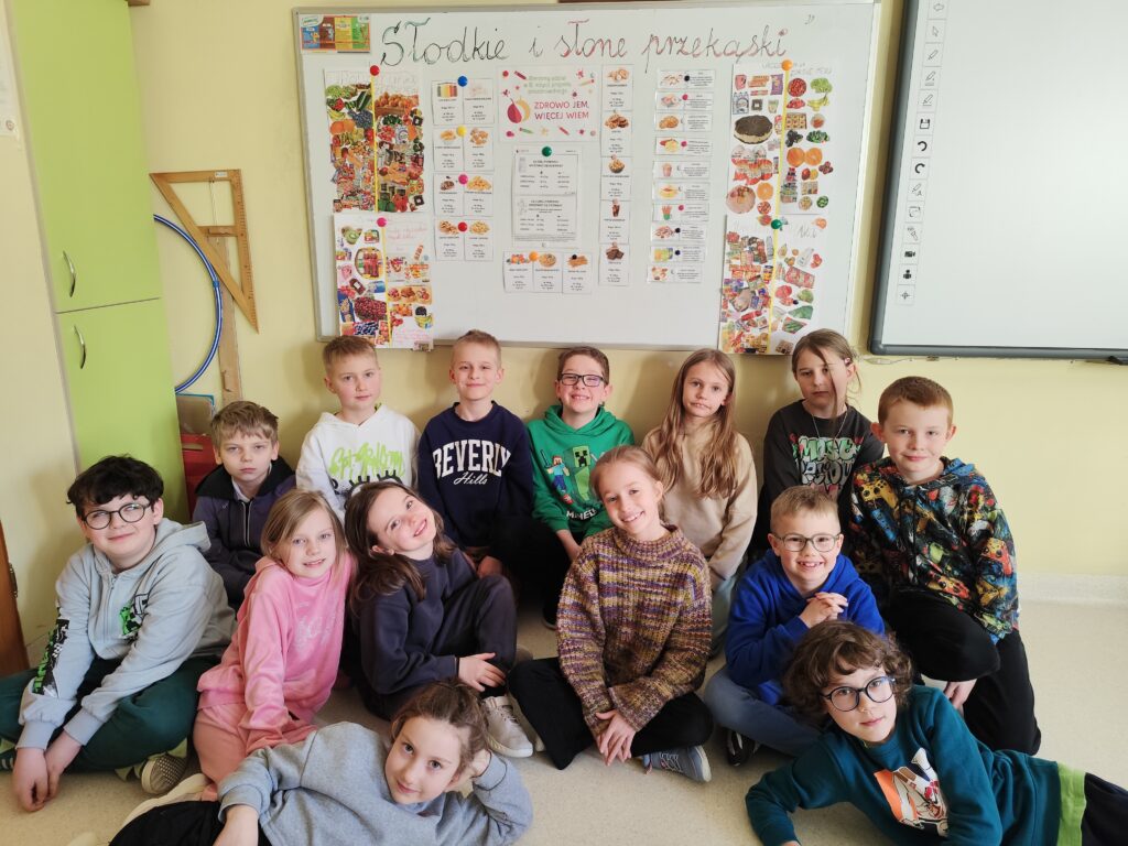 Na zdjęciu znajduje się grupa dzieci siedzących na podłodze w klasie szkolnej. W tle widać tablicę z kolorowym plakatem dotyczącym zdrowego odżywiania, zatytułowanym "Słodkie i słone przekąski". Dzieci są zadowolone, ubrane są w wygodne, codzienne ubrania, niektóre w kolorowe bluzy z nadrukami. Klasa ma jasne ściany, zielone szafki i białą tablicę interaktywną.
