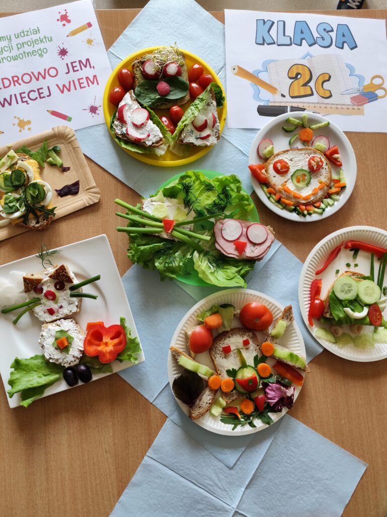 Na zdjęciu widzimy kolorowe i kreatywne kanapki przygotowane z warzyw, pieczywa i nabiału, które ułożono na talerzach. Całość jest częścią projektu edukacyjnego o zdrowym odżywianiu, co potwierdza kartka z napisem „ZDROWO JEM, WIĘCEJ WIEM”. Niektóre kompozycje przypominają uśmiechnięte buzie lub zwierzęta. Na stole leży także kartka z napisem „KLASA 2c.