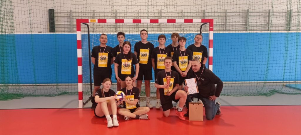 Zdjęcie przedstawia drużynę siatkarską, prawdopodobnie młodzieżową, po meczu lub turnieju. Zawodnicy i trener są ubrani w czarne koszulki z napisem „2004”. Dwóch zawodników siedzi z przodu, trzymając puchar i dyplom. Pozostali stoją za nimi, uśmiechając się do kamery. Na zdjęciu widoczny jest również trener, który trzyma dyplom. Tło stanowi hala sportowa z siatką do siatkówki. Atmosfera jest radosna i triumfalna, sugerująca zwycięstwo w zawodach. Zdjęcie jest ostre i dobrze naświetlone, co pozwala na wyraźne rozpoznanie szczegółów.