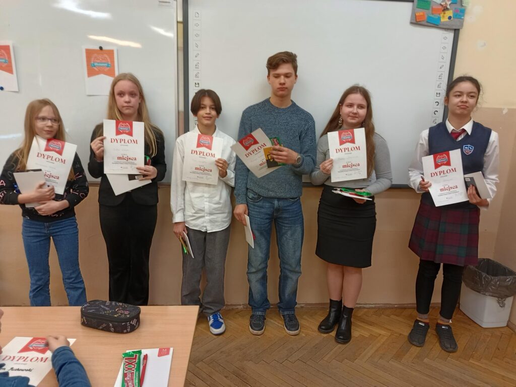Na zdjęciu sześć osób stoi na tle tablicy, trzymając dyplomy i nagrody. Są to zarówno dziewczęta, jak i chłopcy, ubrani w różne stroje – od eleganckich po bardziej codzienne. Każdy z nich trzyma w ręku dyplom z napisem „DYPLOM” oraz słodycze czy zeszyty.
