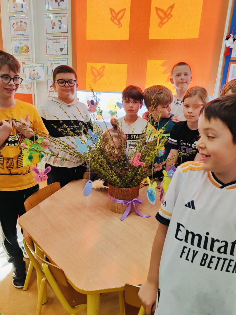 Na zdjęciu znajduje się grupa dzieci stojących wokół stołu w sali przedszkolnej lub szkolnej. Na stole stoi wazon z gałązkami udekorowanymi kolorowymi ozdobami wielkanocnymi, takimi jak jajka i króliczki. W tle widać kolorową roletę z wyciętymi motywami wiosennymi oraz plakaty edukacyjne na ścianie. Dzieci wyglądają na zaangażowane i zadowolone, prawdopodobnie uczestniczyły w tworzeniu tych dekoracji.