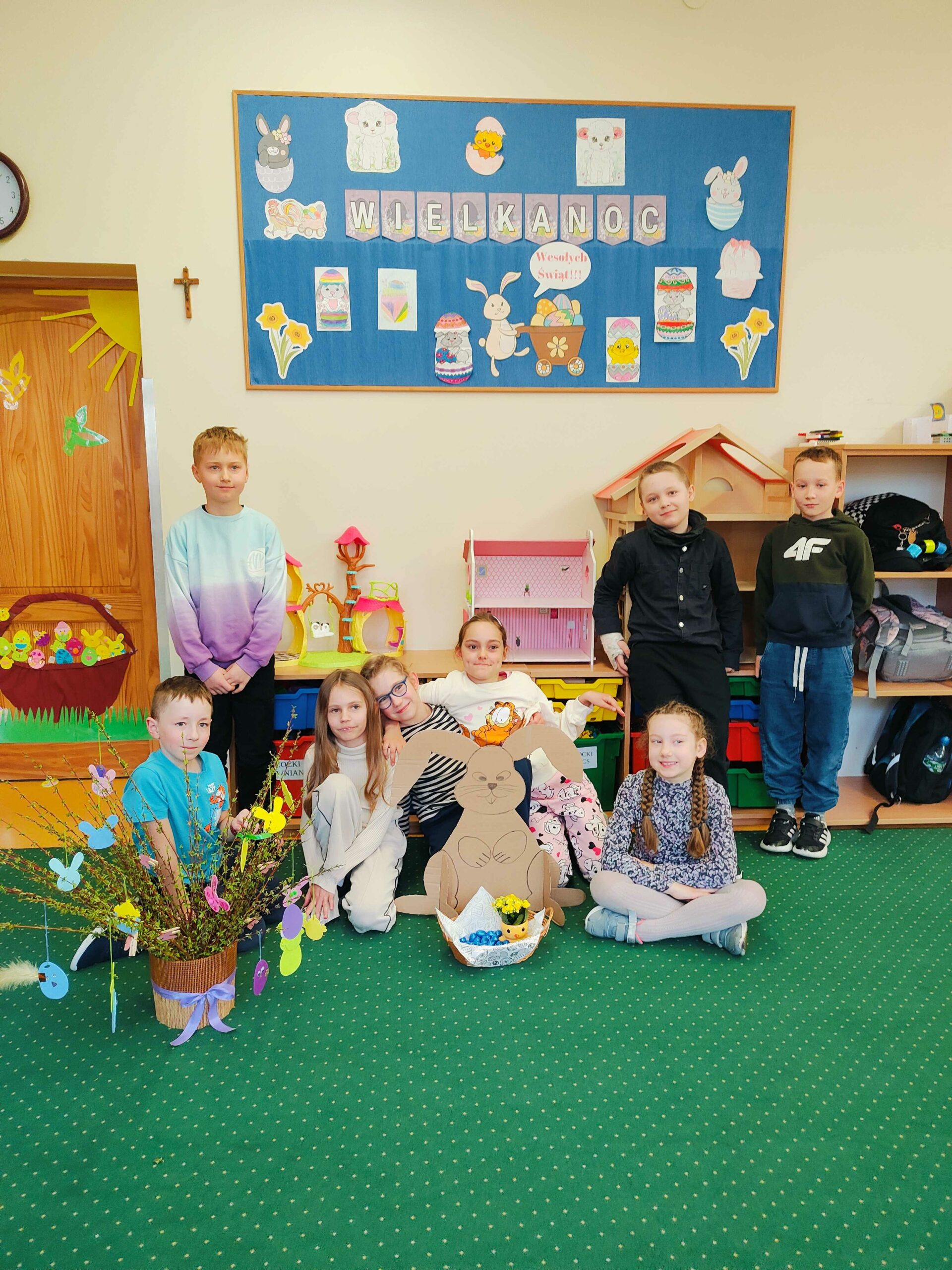 Na zdjęciu znajduje się grupa ośmiorga dzieci w klasie szkolnej, pozujących przed wielkanocną dekoracją. Na tablicy widnieje napis „Wielkanoc” oraz kolorowe rysunki z motywami świątecznymi, takimi jak króliczki, jajka i kwiaty. Dzieci ubrane są w codzienne ubrania i wyglądają na radosne, niektóre z nich siedzą na dywanie obok wielkanocnego koszyczka i dużego kartonowego zajączka. W tle widać półki z zabawkami, domek dla lalek oraz wielkanocny stroik z gałązkami ozdobionymi wycinankami.