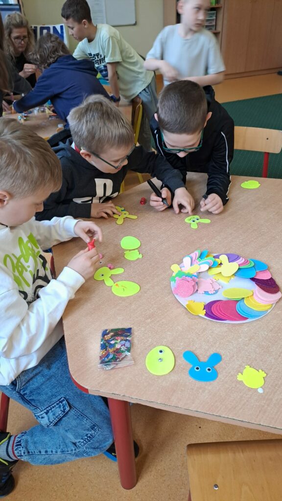 Na zdjęciu widać dzieci pracujące przy stole nad wielkanocnymi ozdobami. Chłopcy skupieni są na wycinaniu i ozdabianiu kolorowych kształtów z papieru, takich jak zajączki, kurczaczki i jajka. Na stole leży wiele barwnych elementów i ozdób, m.in. cekiny i gotowe wycinanki z pianki. W tle inni uczniowie oraz nauczycielka również angażują się w kreatywne zajęcia, co tworzy atmosferę skupienia i zabawy.