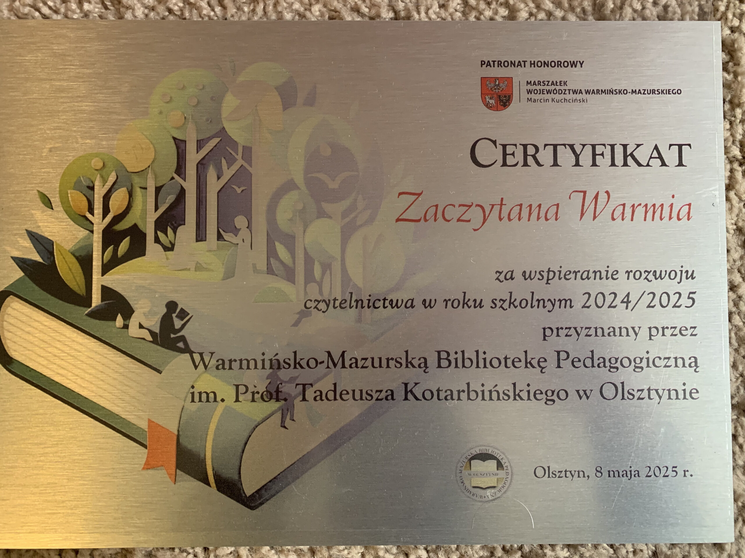 po prawej stronie widoczny jest napis CERTYFIKAT Zaczytana Warmia za wsparcie rozwoju czytelnictwa w roku szkolnym 2024/2025 przyznany przez Warmińsko-Mazurską Bibliotekę Pedagogiczną im. Prof. Tadeusza Kotarbińskiego w Olsztynie. Po lewej stronie widoczny jest rysunek leżącej książki, a z niej wyrastające drzewa