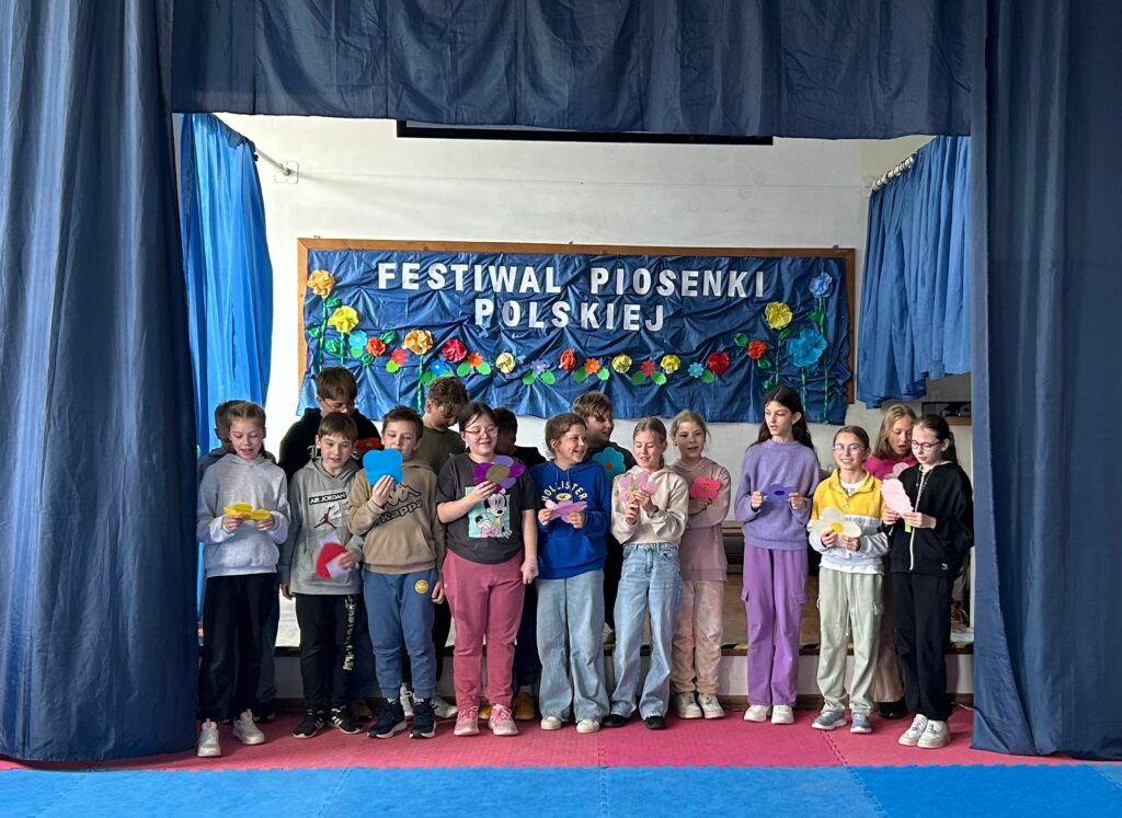 Uczniowie klasy 4a śpiewają piosenkę. W ręku trzymają kolorowe kartki/kwiatki z tekstem. Są uśmiechnięci. Stoją na scenie, po obu stronach wiszą granatowe zasłony, w tle widnieje tablica z napisem Festiwal Piosenki Polskiej i kolorowymi kwiatami.
