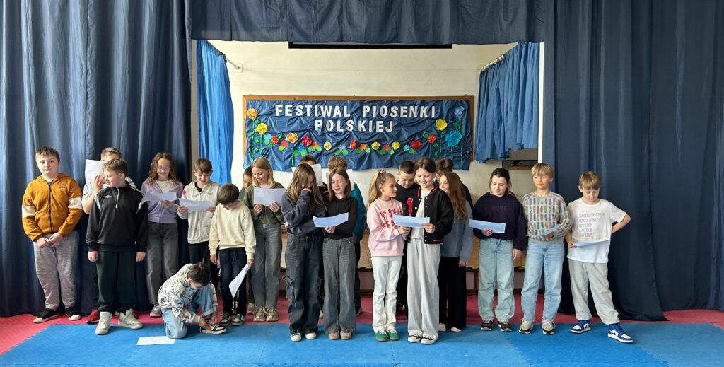 Uczniowie klasy 4b śpiewają piosenkę. W ręku trzymają białe kartki z tekstem. Niektórzy są uśmiechnięci lub skupieni. Stoją na scenie, po obu stronach wiszą granatowe zasłony, w tle widnieje tablica z napisem Festiwal Piosenki Polskiej i kolorowymi kwiatami.