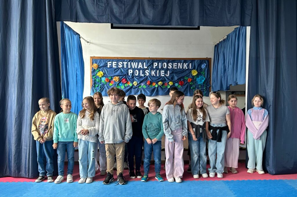 Uczniowie klasy 4c śpiewają piosenkę. Jedni są uśmiechnięci, inni skupieni na aktywności. Stoją na scenie, po obu stronach wiszą granatowe zasłony, w tle widnieje tablica z napisem Festiwal Piosenki Polskiej i kolorowymi kwiatami.