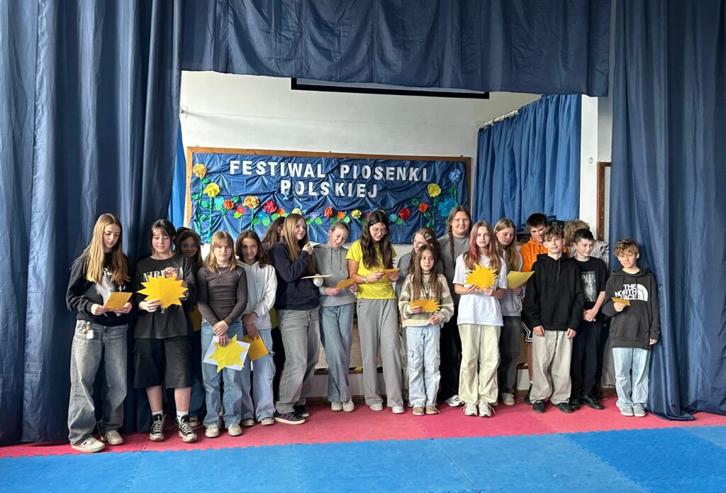 Uczniowie klasy 6a śpiewają piosenkę. W ręku trzymają żółte kartki/słoneczka z tekstem. Są uśmiechnięci. Stoją na scenie, po obu stronach wiszą granatowe zasłony, w tle widnieje tablica z napisem Festiwal Piosenki Polskiej i kolorowymi kwiatami.