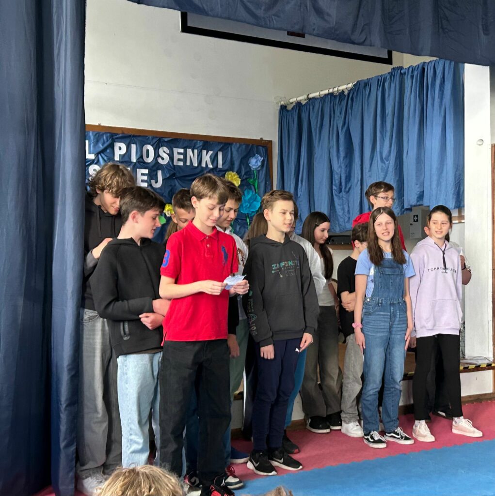 Uczniowie klasy 6c śpiewają piosenkę. Niektórzy trzymają w ręku kartkę z tekstem. Jedni są uśmiechnięci, inni skupieni na aktywności. Stoją na scenie, po obu stronach wiszą granatowe zasłony, w tle widnieje tablica z napisem Festiwal Piosenki Polskiej i kolorowymi kwiatami.