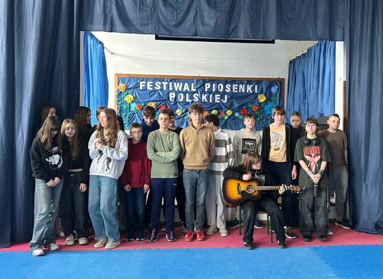 Uczniowie klasy 7a śpiewają piosenkę. Są skupieni na aktywności. Stoją na scenie, po obu stronach wiszą granatowe zasłony, w tle widnieje tablica z napisem Festiwal Piosenki Polskiej i kolorowymi kwiatami. Jedna dziewczynka siedzi na krześle i akompaniuje na gitarze.