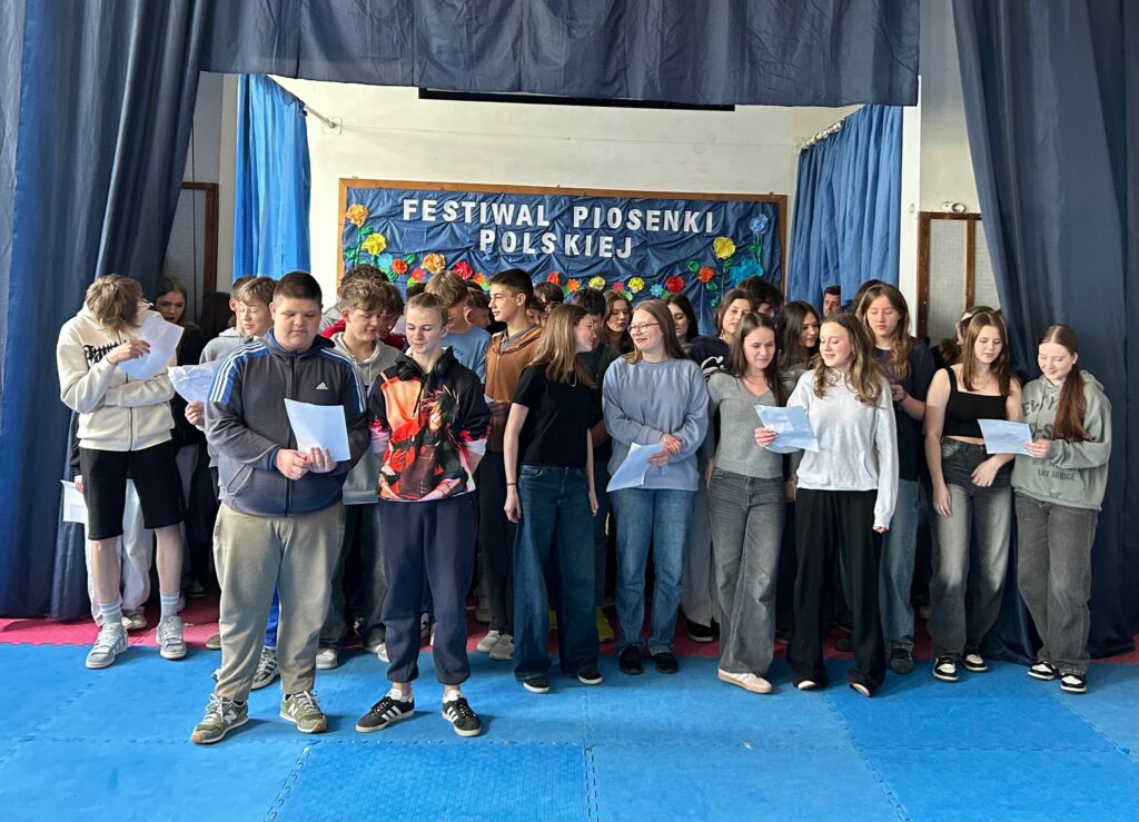 Uczniowie dwóch klas 8a i 8b śpiewają piosenkę. Niektórzy trzymają w ręku kartkę z tekstem. Jedni są uśmiechnięci, inni skupieni na aktywności. Stoją na scenie, po obu stronach wiszą granatowe zasłony, w tle widnieje tablica z napisem Festiwal Piosenki Polskiej i kolorowymi kwiatami.
