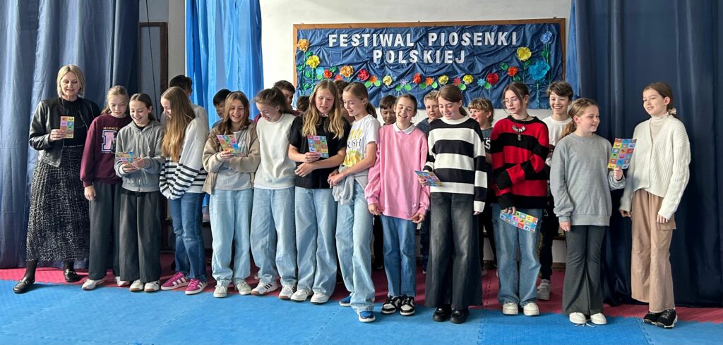 Uczniowie klasy 5a z wychowawczynią śpiewają piosenkę. Niektórzy trzymają w ręku kolorową kartkę z tekstem. Jedni są uśmiechnięci, inni skupieni na aktywności. Stoją na scenie w dwóch rzędach, po obu stronach wiszą granatowe zasłony, w tle widnieje tablica z napisem Festiwal Piosenki Polskiej i kolorowymi kwiatami.